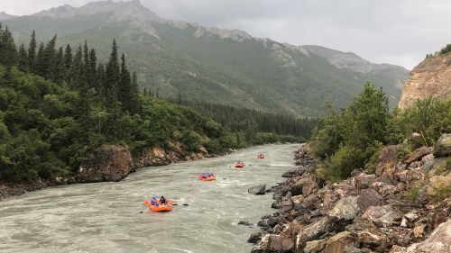 A group of people in orange rafts navigating a river surrounded by lush green mountains and rocky banks.