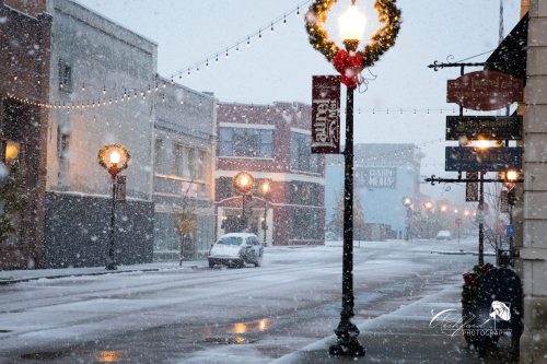 A snowy street scene with decorated lampposts and empty storefronts, creating a quiet winter atmosphere.