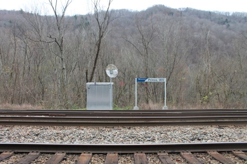 A train station sign for Prince, WV, near railway tracks, with a backdrop of bare trees and a distant mountain.