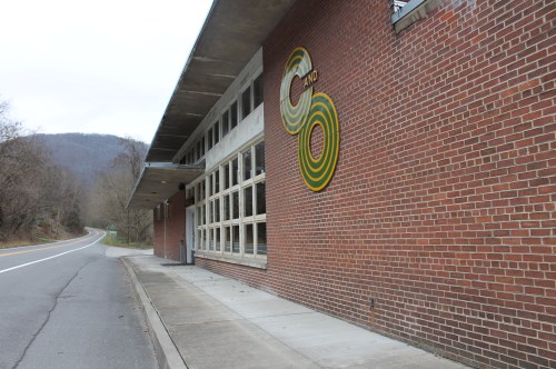 A brick building with large circular yellow and green designs, located near a road and surrounded by trees and hills.