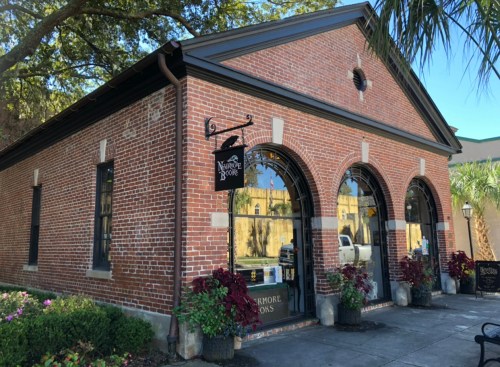 A brick building with large arched windows and a sign, surrounded by greenery and colorful plants.