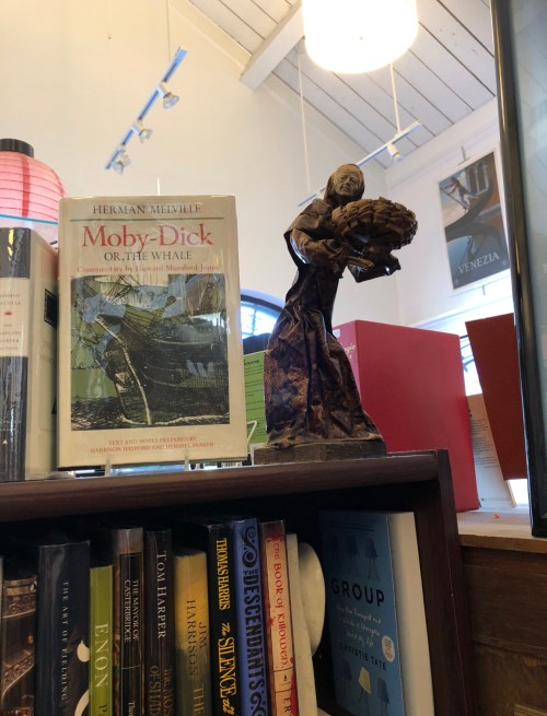 A book titled "Moby-Dick" beside a small statue of a woman holding a basket, displayed on a wooden shelf.