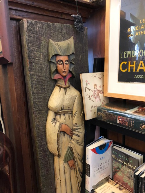 A carved wooden figure of a woman in a white dress and bonnet, displayed among books on a shelf.