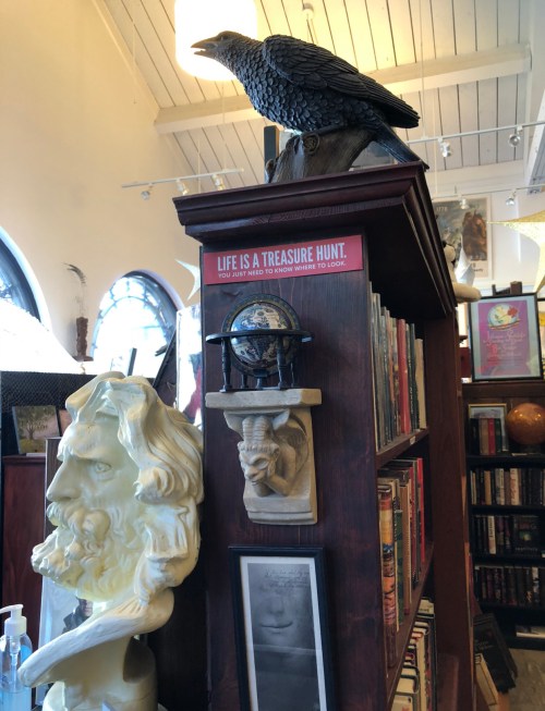 A wooden bookshelf with a raven statue, a globe, and a bust, featuring a sign that reads "Life is a treasure hunt."