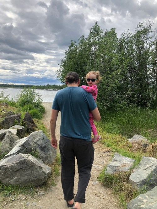 A person carries a child on their shoulder along a rocky path by a river, surrounded by greenery and cloudy skies.