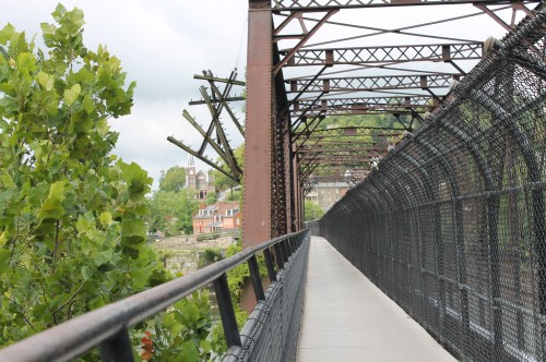 A metal bridge with a fenced walkway, surrounded by greenery and buildings in the background under a cloudy sky.