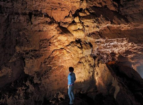 A person stands in a cave, illuminated by warm light, showcasing textured rock formations on the walls.