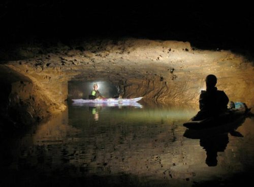 Two kayakers explore a dark cave, illuminated by their lights reflecting on the calm water.