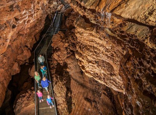 Visitors explore a cave on a metal walkway, surrounded by textured rock formations and a small waterfall.