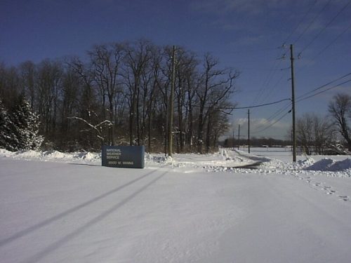 Snow-covered landscape with a sign for the National Vehicle Center and bare trees in the background under a clear blue sky.