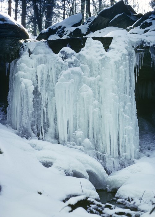 A frozen waterfall surrounded by snow-covered rocks and trees in a winter landscape.