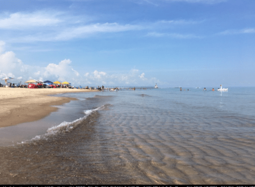 A sunny beach scene with gentle waves, colorful umbrellas, and people enjoying the water near a pier.