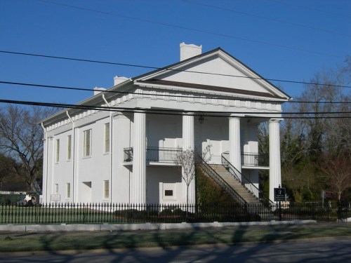 A white, classical-style building with columns and a staircase, set against a clear blue sky.