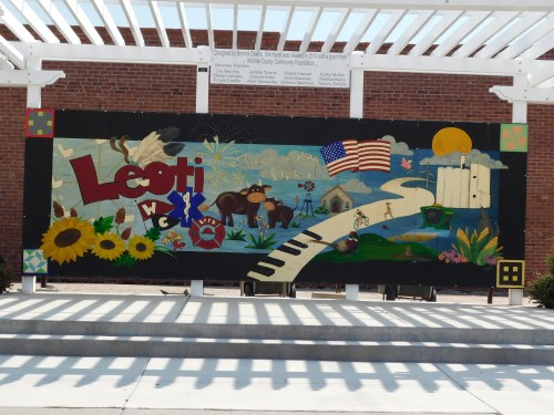Colorful mural featuring sunflowers, a piano, an American flag, and various community symbols against a brick wall.