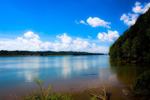 A serene river scene with clear blue skies, fluffy clouds, and lush greenery along the water's edge.