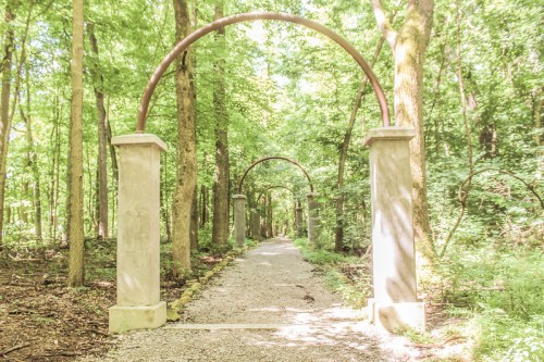 A serene forest path lined with stone pillars and arches, surrounded by lush green trees.
