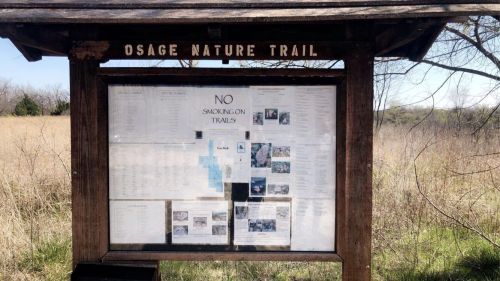 A wooden signboard displaying information about the Osage Nature Trail, with rules and maps, surrounded by tall grass.