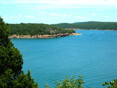 A serene lake surrounded by green hills and trees under a clear blue sky. Gentle ripples on the water's surface.