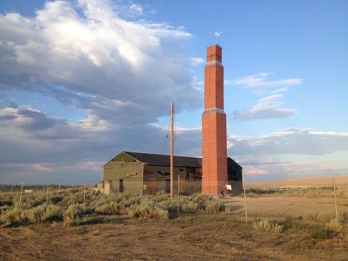 A tall brick chimney next to a green building, surrounded by sparse vegetation and under a cloudy sky.