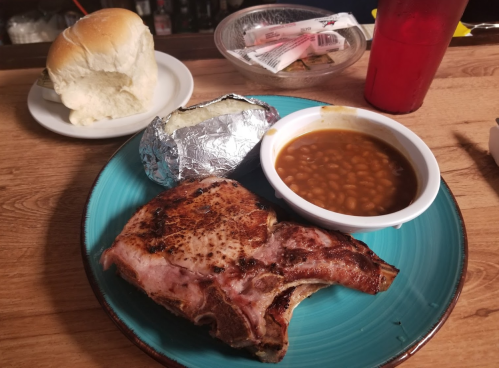 A plate with a grilled pork chop, baked potato, baked beans, and a roll, with a drink in the background.