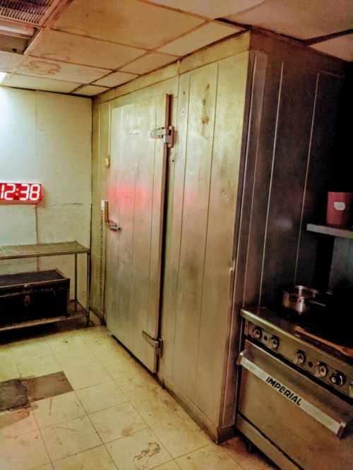 A dimly lit kitchen with a large metal fridge, stove, and a digital clock displaying 12:38 on the wall.