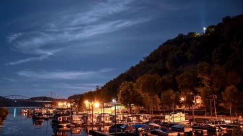 Nighttime view of a marina with boats, illuminated by lights, against a backdrop of a hill and starry sky.
