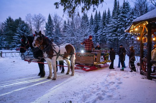 A horse-drawn sleigh in a snowy landscape, with people gathered around a cozy cabin decorated with lights.