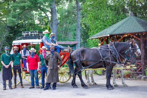 A group of people in masks stands by a horse-drawn carriage in a wooded area, with a gazebo in the background.