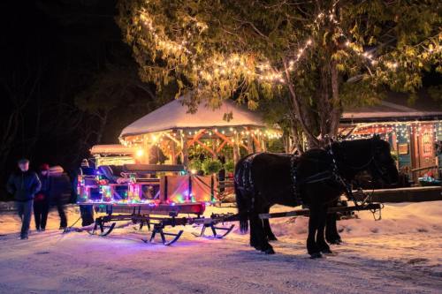 A festive horse-drawn sleigh adorned with lights, parked near a warmly lit cabin in a snowy landscape at night.