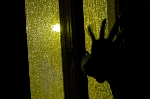 A shadowy figure presses against a rain-soaked window, illuminated by a warm light outside.