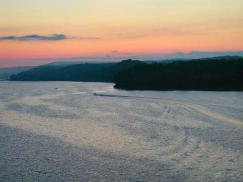 A serene sunset over a calm river, with silhouettes of hills and a boat leaving a wake in the water.