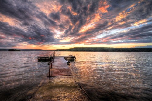 A wooden dock extends into a calm lake, under a dramatic sky filled with colorful clouds at sunset.