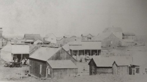Black and white photo of a small settlement with wooden houses and a few people visible outside.