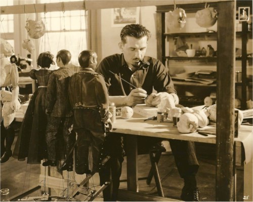 A man works on puppet heads at a table, surrounded by hanging puppets in a well-lit workshop.
