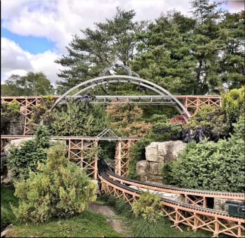 A miniature train track winds through lush greenery and rocky terrain, featuring an arch bridge overhead.