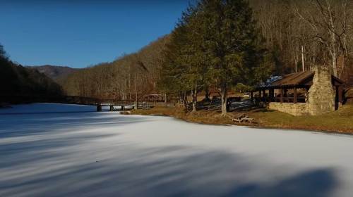 A serene winter scene of a frozen lake surrounded by trees and a rustic pavilion in the background.