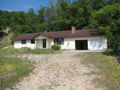 A single-story house with a brown roof, surrounded by greenery and a gravel driveway.