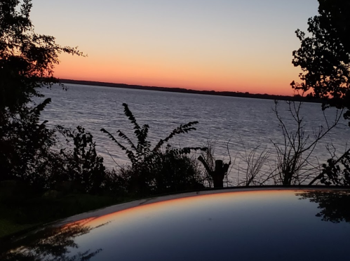 A serene sunset over a calm lake, with silhouettes of trees and a car roof reflecting the colorful sky.
