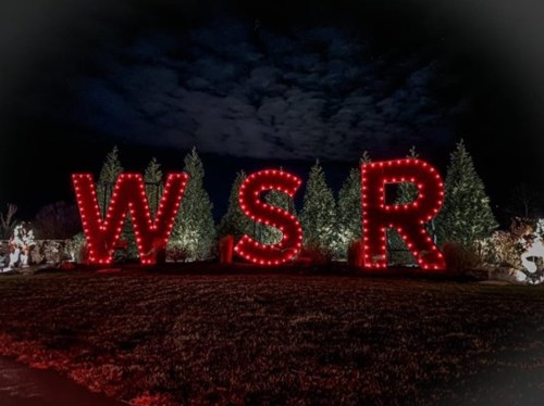 Large illuminated letters "W," "S," and "R" in red lights, surrounded by evergreen trees at night.