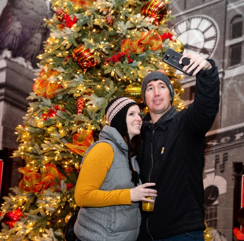 A couple takes a selfie in front of a beautifully decorated Christmas tree with lights and ornaments.