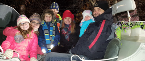 A group of seven people, including children and adults, smiling together in a festive outdoor setting.