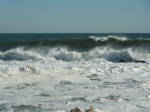Waves crashing on a rocky shore under a clear blue sky, creating white foam on the water's surface.