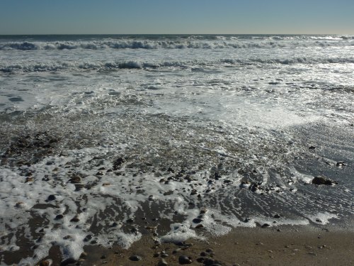 Waves gently crash on a sandy beach, creating foam and ripples under a clear blue sky.