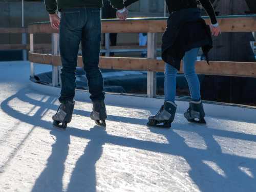 A couple ice skating hand in hand, casting long shadows on the rink.