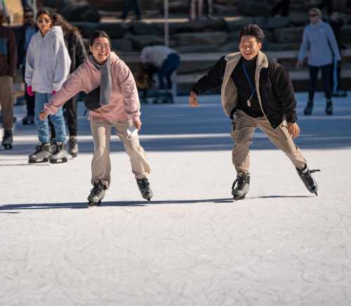 Two people ice skating joyfully on a rink, surrounded by others enjoying the activity.