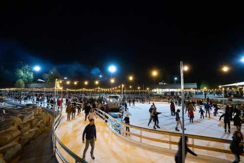 A lively outdoor ice skating rink at night, filled with skaters and illuminated by string lights and bright overhead lights.