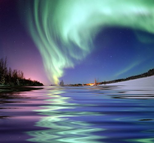 A stunning view of the Northern Lights reflecting on a calm river, with trees and a distant warm glow on the shore.