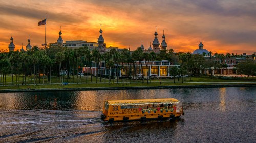 A yellow boat glides on water at sunset, with palm trees and historic buildings silhouetted against a colorful sky.
