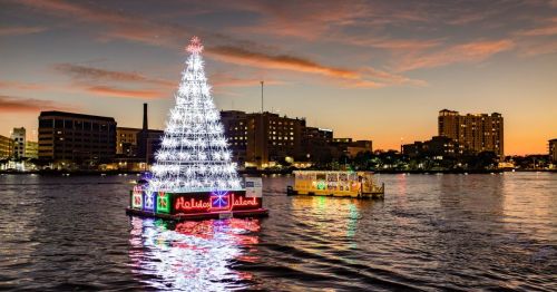 A brightly lit Christmas tree on a floating platform in a river at sunset, with city buildings in the background.
