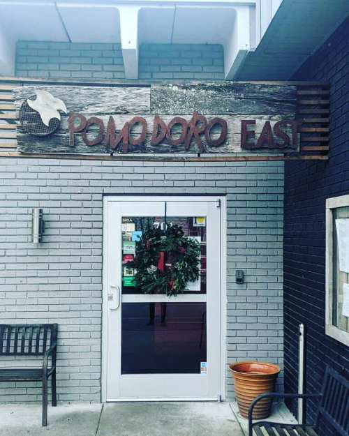 Sign for Pomodoro East above a door, with a wreath on the door and benches nearby.